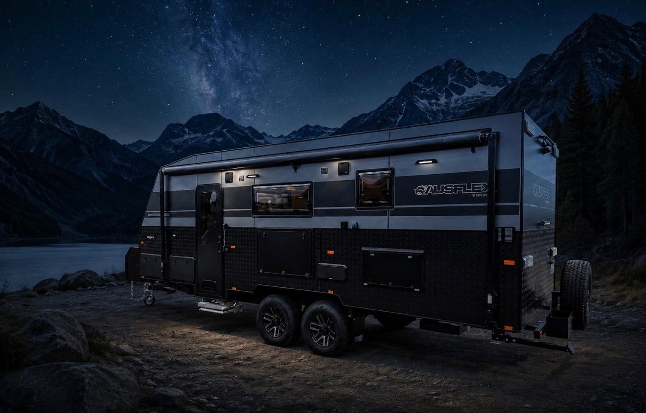 An Ausflex caravan parked beside a lake at night, awning out and lights on, under a star-filled sky with mountains behind