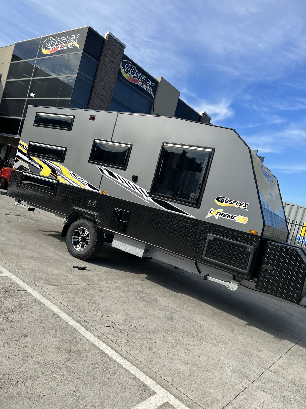 Ausflex Xtreme 18 in charcoal with yellow graphics, front three-quarter view outside the showroom